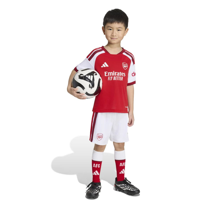 A young boy modeling a red and white Arsenal soccer kit, holding a soccer ball against a clean white background.