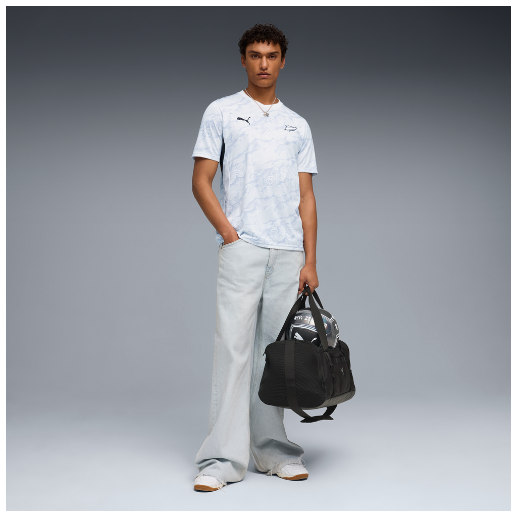 Person wearing white NZ All Whites Football Jersey holding a duffel bag and football, wearing jeans and sneakers