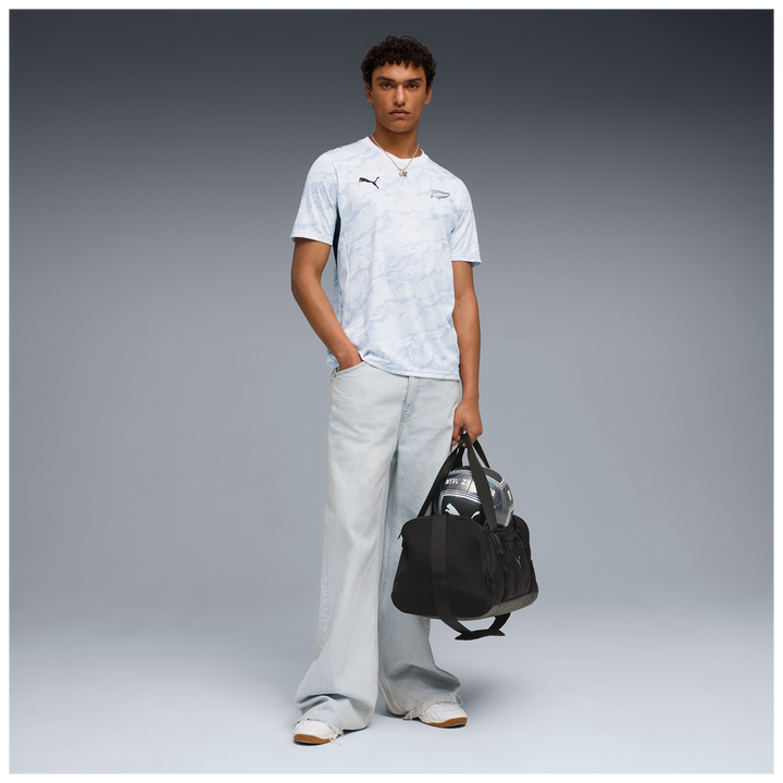 Person wearing white NZ All Whites Football Jersey holding a duffel bag and football, wearing jeans and sneakers