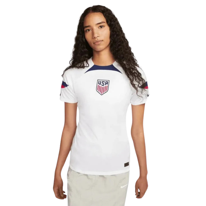 A woman modeling a white Nike USA soccer jersey with navy and red accents, featuring the USA crest on the chest.